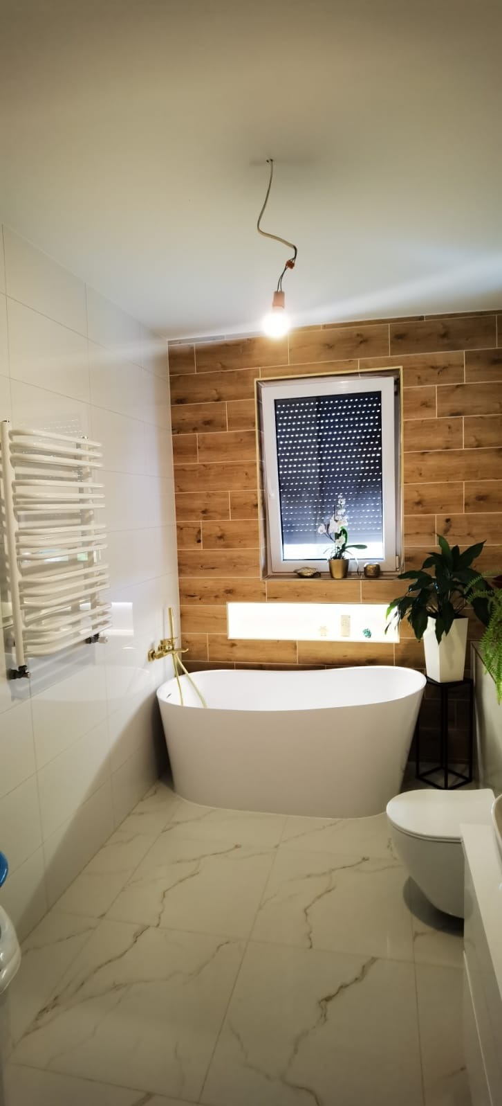 Nowoczesna łazienka z wolnostojącą wanną, drewnopodobnymi płytkami na ścianie i marmurową podłogą. Minimalistyczny grzejnik drabinkowy i złota armatura.