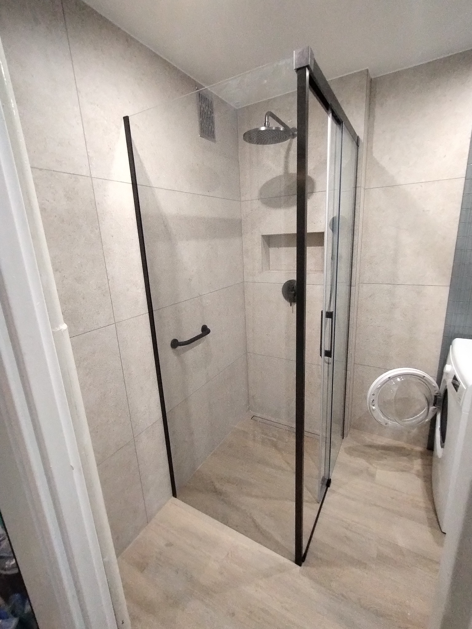 Nowoczesna kabina prysznicowa z czarnymi profilami i deszczownicą w wykończonej łazience z beżowymi płytkami i pralką. Minimalistyczny design.