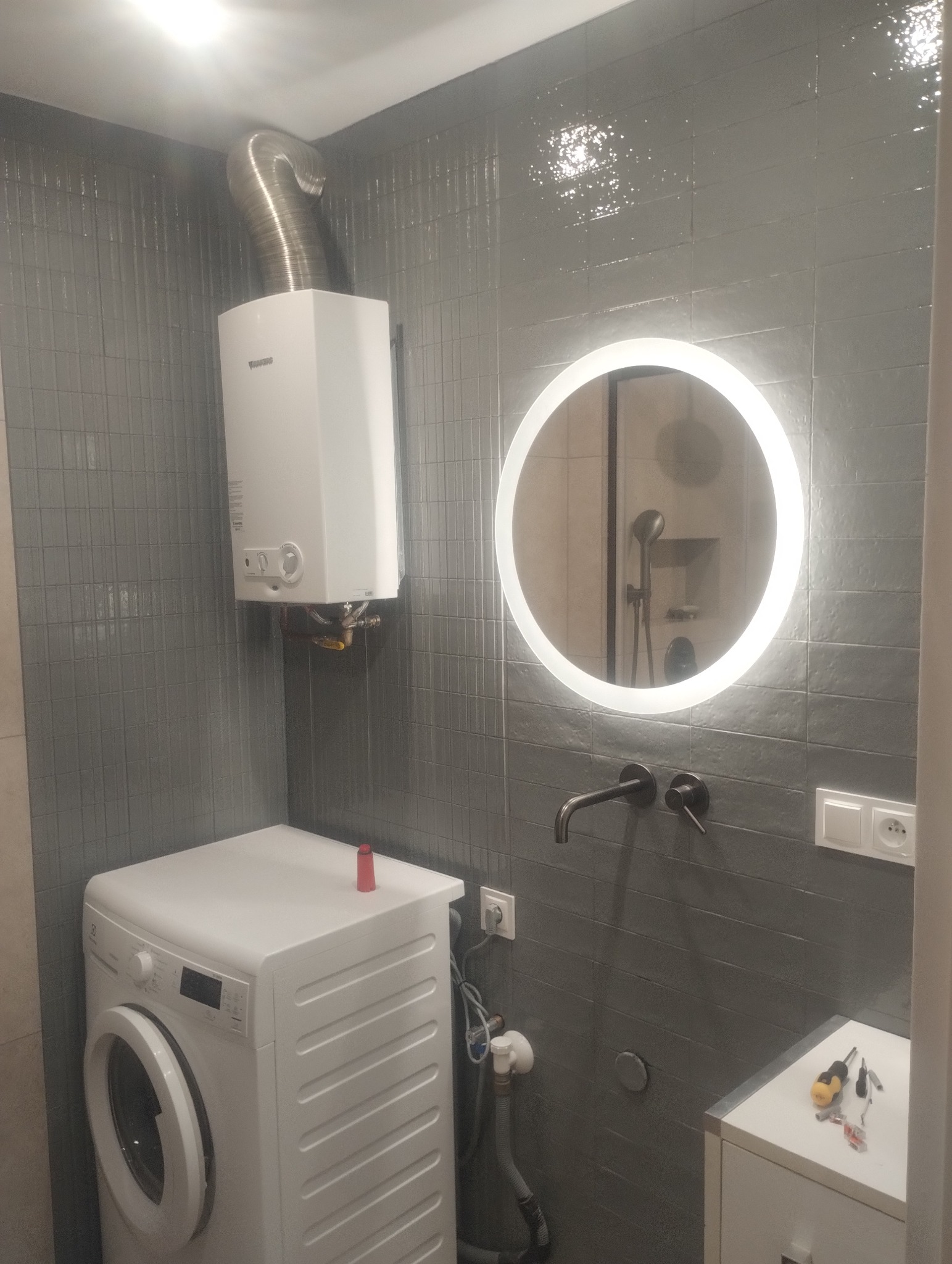 Szara łazienka po remoncie: podświetlane okrągłe lustro, pralka, bojler z rurą odprowadzającą spaliny, ciemna armatura i płytki imitujące cegłę.