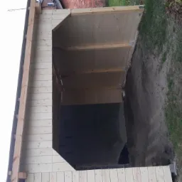 Surowy stan rozbudowy domu: widok na szkielet drewnianej konstrukcji z betonową wylewką w środku, otoczony ziemią i trawą.