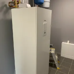 Biała pompa ciepła Panasonic Aquarea z widocznymi niebieskimi zbiornikami wyrównawczymi i miedzianą instalacją hydrauliczną na szarej ścianie, drabina monterska obok urządzenia.