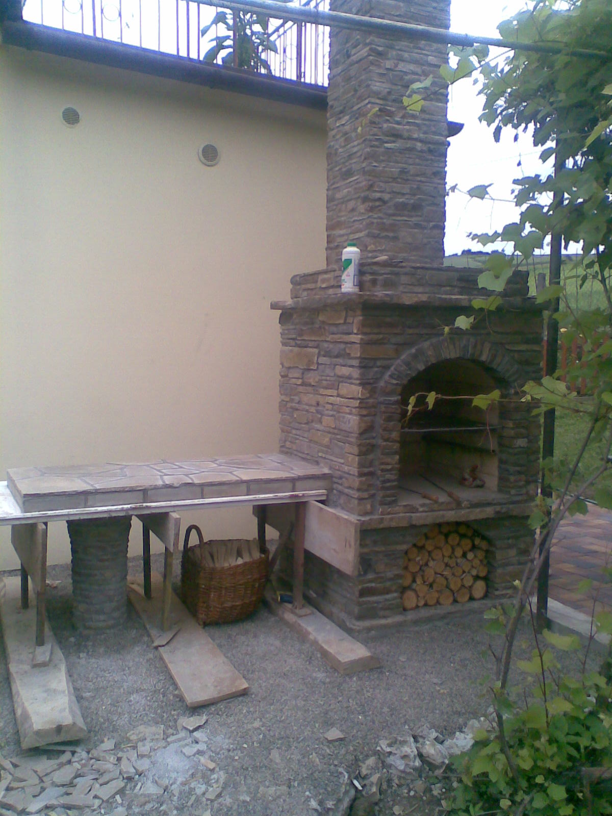 Murowany grill ogrodowy z kamienia, z miejscem na drewno i blatem roboczym w trakcie budowy, widoczna butelka płynu na górze konstrukcji.