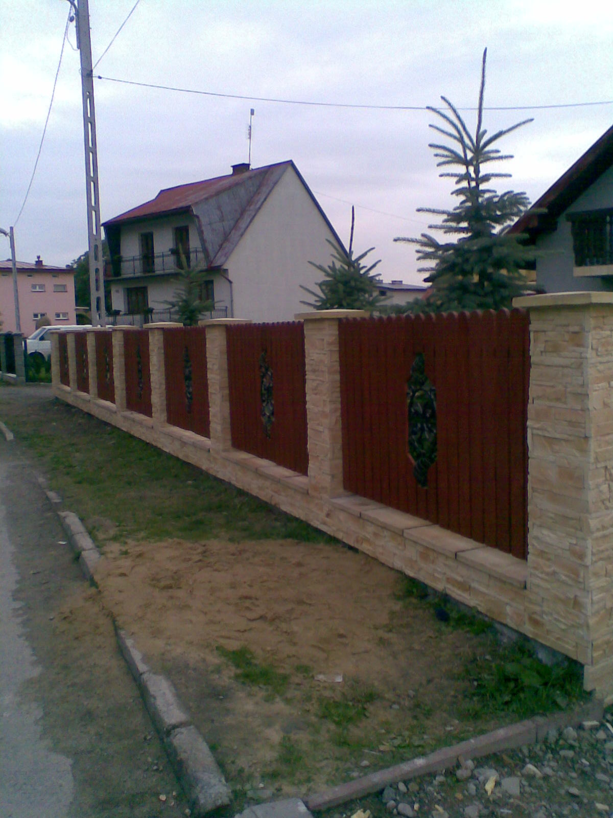 Nowoczesne ogrodzenie drewniane z elementami dekoracyjnymi i kamiennymi słupkami, widok z poziomu ulicy, w tle domy jednorodzinne.