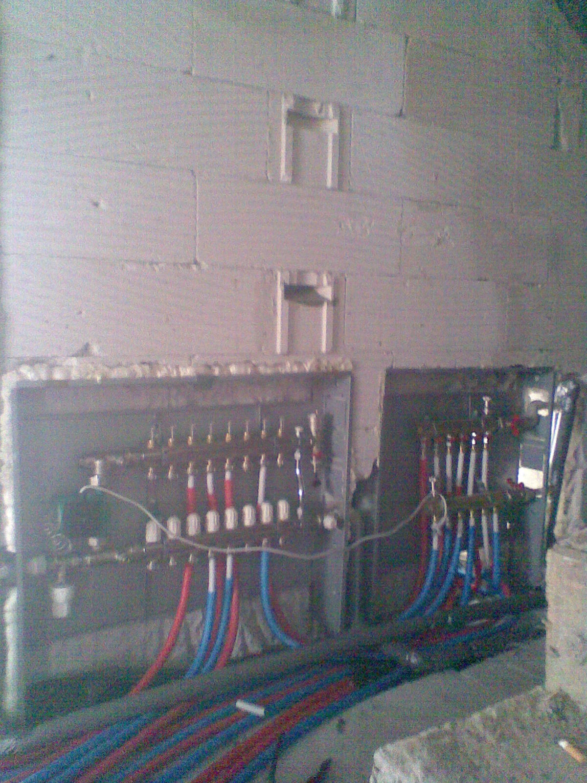 Rozdzielacze ogrzewania podłogowego zamontowane w ścianie z betonu komórkowego, widoczne rury w kolorze niebieskim i czerwonym.