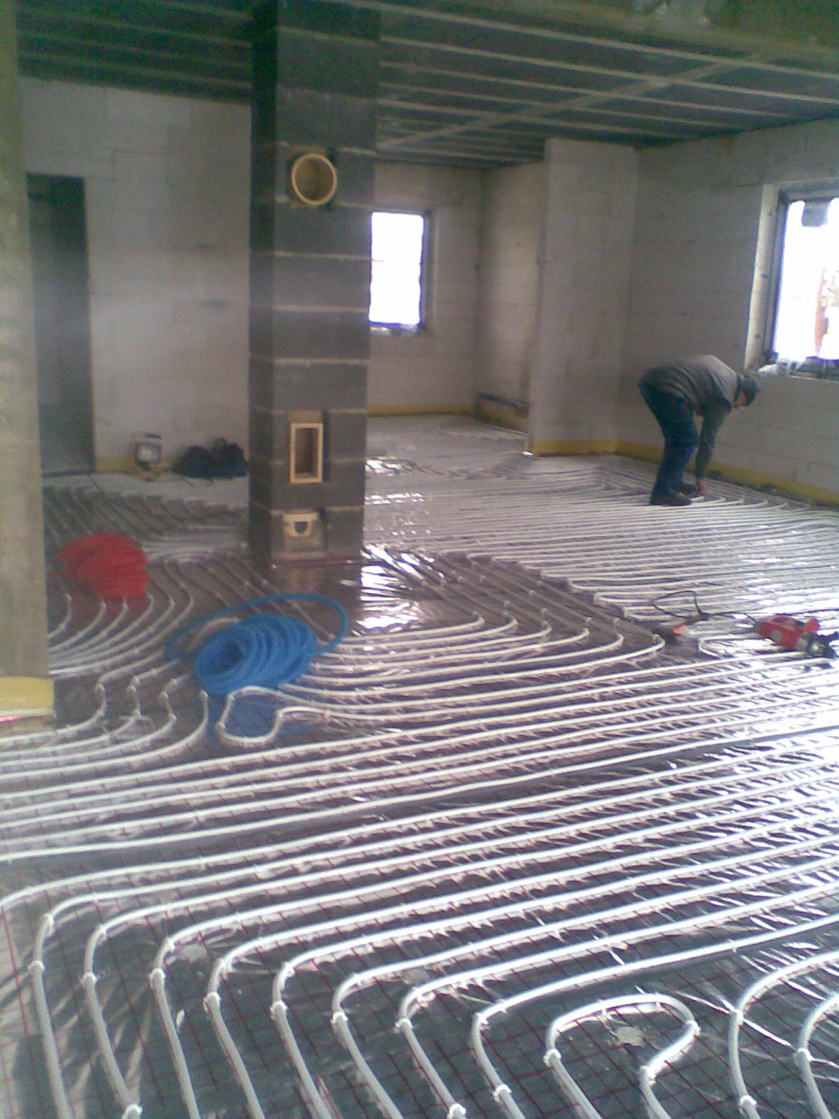 Instalacja ogrzewania podłogowego w surowym pomieszczeniu, widoczne rury grzewcze ułożone na siatce, zwinięte zwoje rur w kolorze niebieskim i czerwonym oraz pracownik pochylony nad instalacją przy...