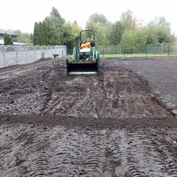 King Grass - Traktor wyrównuje teren pod trawnik, widoczne ślady opon i przygotowany obszar ziemi, w tle ogrodzenie panelowe i drzewa.