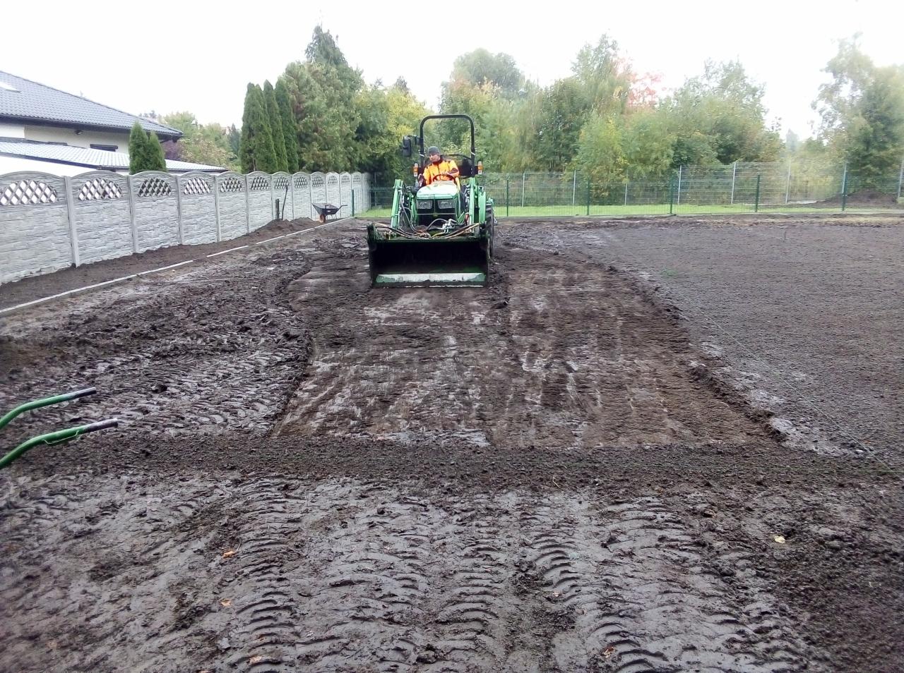 Traktor wyrównuje teren pod trawnik, widoczne ślady opon i przygotowany obszar ziemi, w tle ogrodzenie panelowe i drzewa.