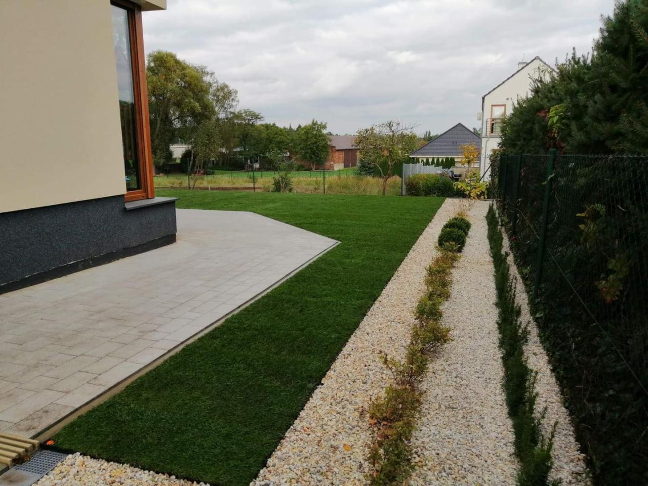 Nowoczesny ogród przydomowy z równo przystrzyżonym trawnikiem graniczącym z kostką brukową, ścieżką wysypaną białym żwirem i niskimi krzewami wzdłuż ogrodzenia.