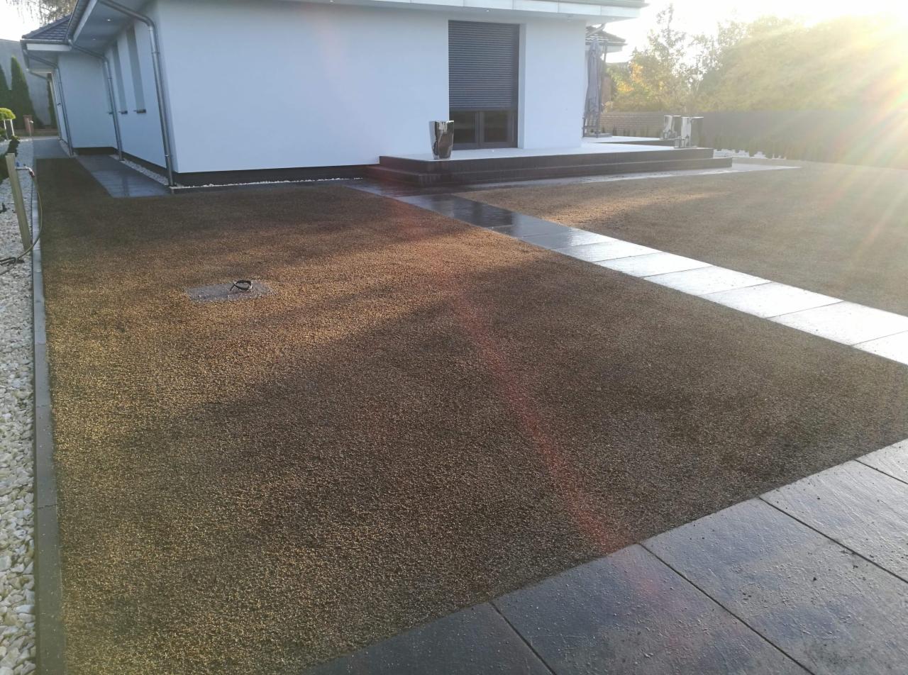 Nowo założony trawnik z widoczną instalacją nawadniającą w postaci okrągłego włazu, obrzeża z jasnych kamyków i ciemnych płyt chodnikowych, dom z białym tynkiem w tle.