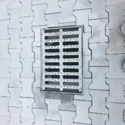 Ułożona szara kostka brukowa typu 'H' z metalową kratką ściekową w centralnej części; widoczne fragmenty obuwia roboczego z czerwonymi sznurówkami.