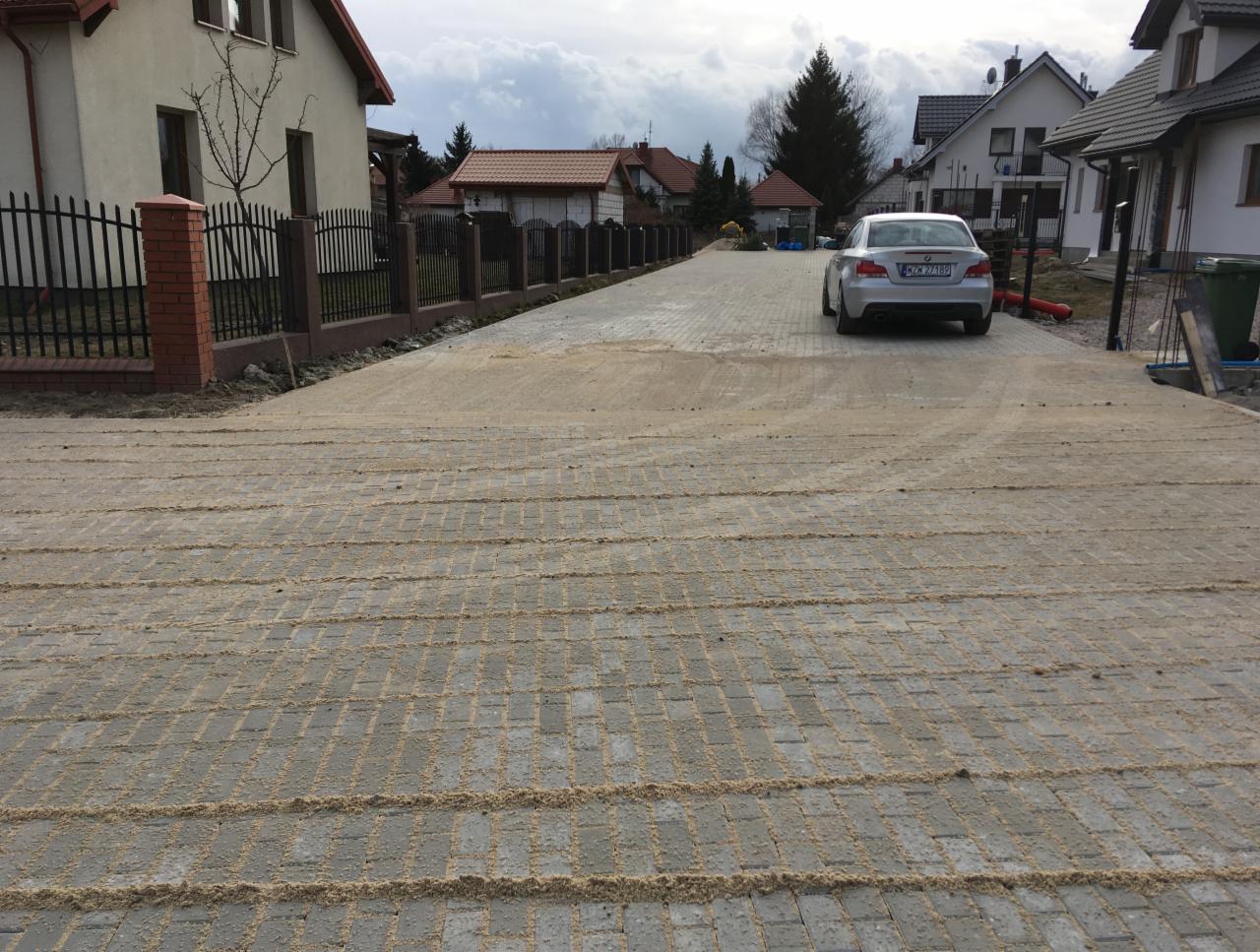 Ułożona kostka brukowa na posesji z widocznymi śladami po zagęszczarce, zasypana piaskiem, w tle domy jednorodzinne i zaparkowany srebrny samochód.