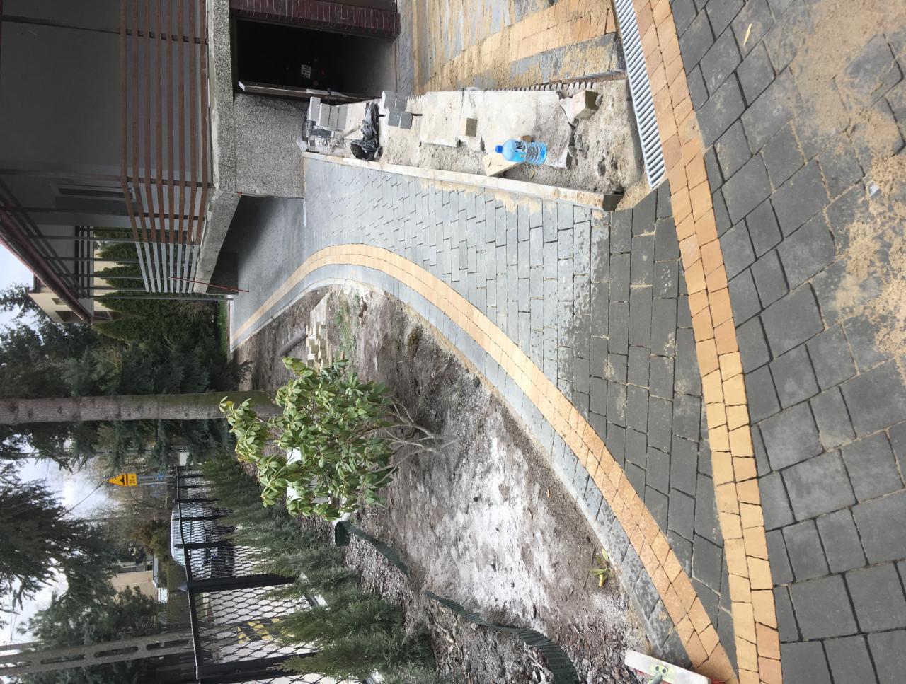 Niedokończona ścieżka z kostki brukowej w odcieniach szarości i beżu, z widocznym korytkiem odwadniającym, obok domu z balkonem i młodymi roślinami.