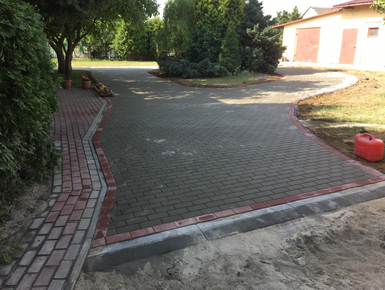 Ułożona ścieżka z szarej kostki brukowej z czerwonym obramowaniem, prowadząca do budynku gospodarczego, obok widoczny czerwony kanister.