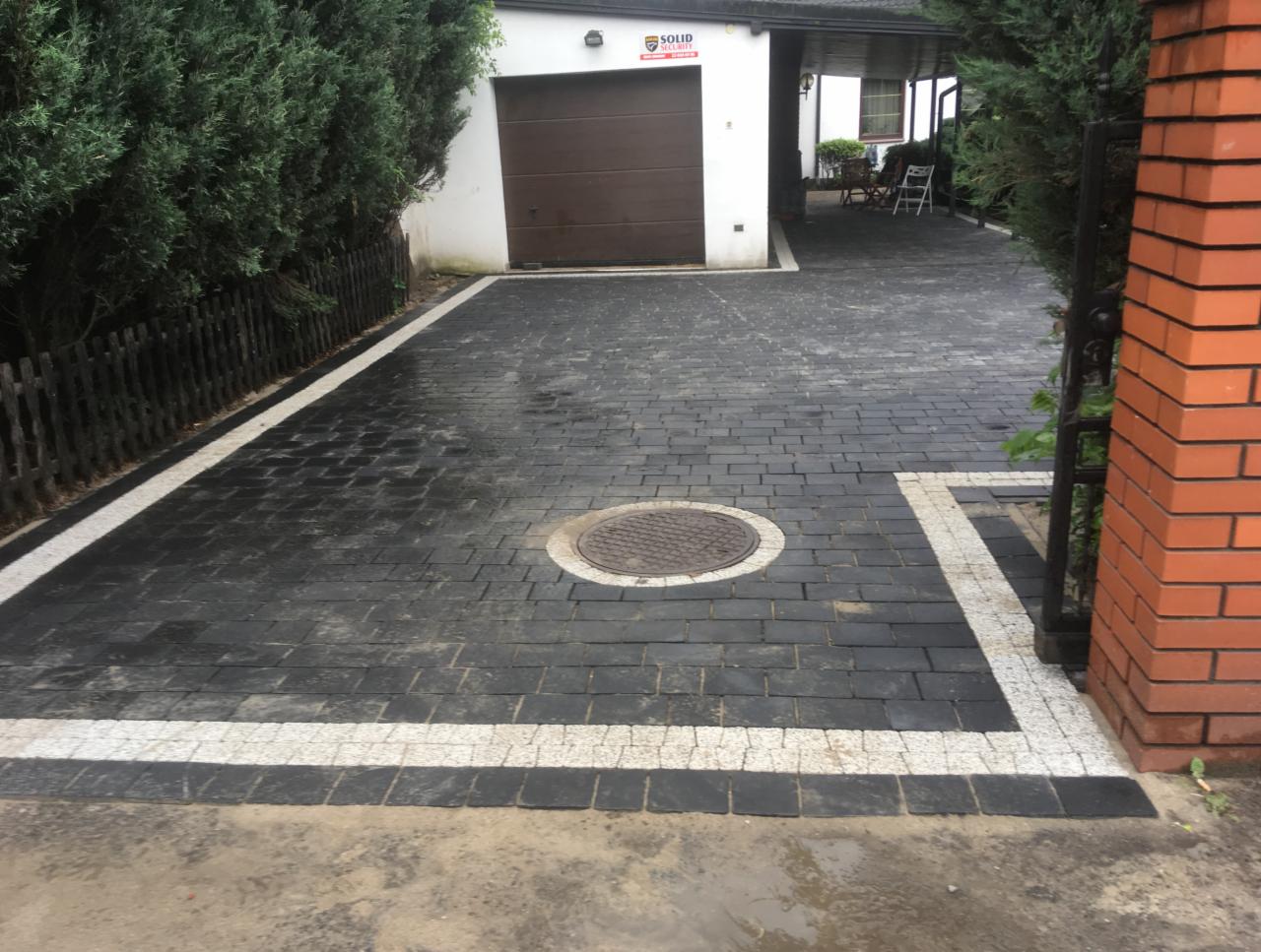 Czarna kostka brukowa z białymi obramowaniami prowadząca do garażu, widoczna studzienka kanalizacyjna i brama z cegły.