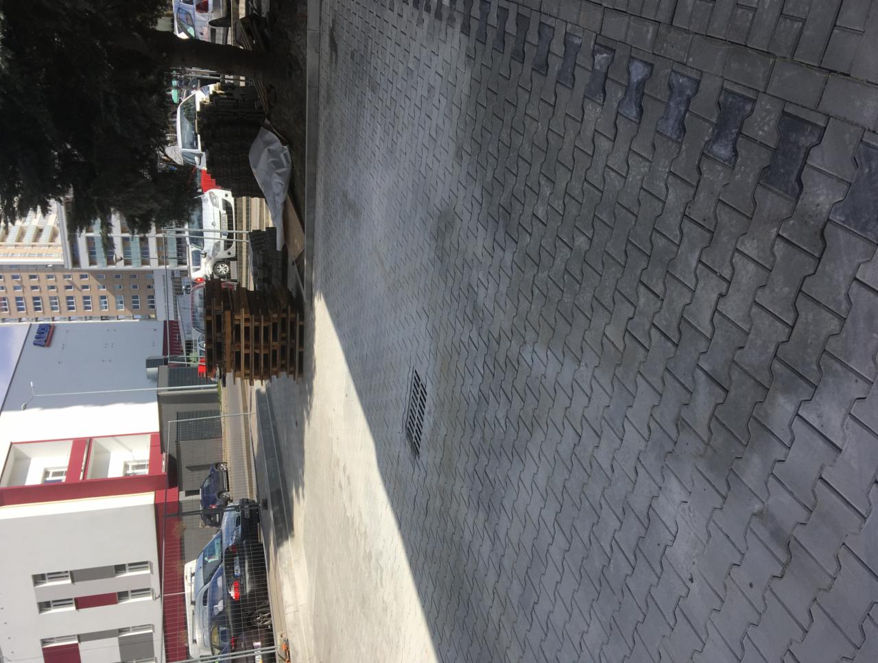 Ułożona szara kostka brukowa typu 'kość', widoczna studzienka kanalizacyjna, paleta z kostką i budynki w tle.