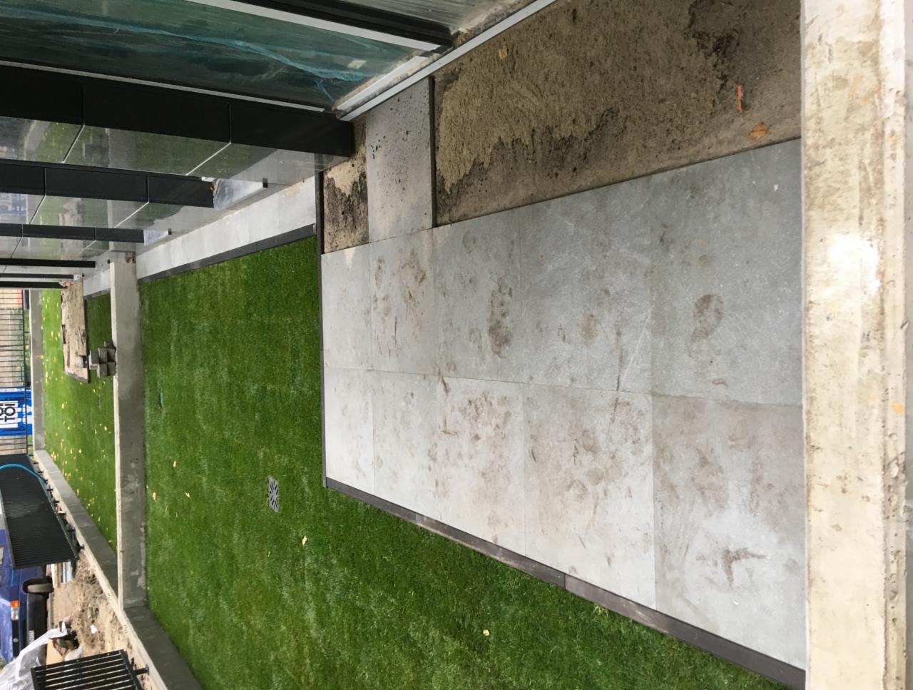 Fragment placu z ułożoną jasnoszarą kostką, sąsiadujący z obszarem pokrytym sztuczną trawą, widoczne zadaszenie i betonowe filary, ślady zabrudzeń na kostce.