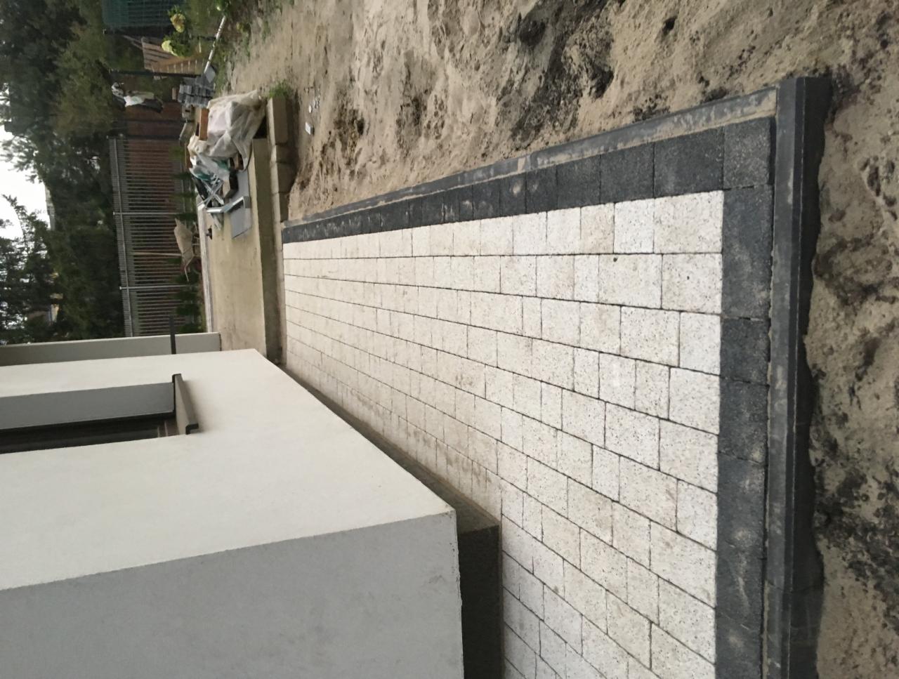 Ułożona kostka brukowa w odcieniach bieli z ciemnoszarą obwódką, widoczna wzdłuż ściany budynku i częściowo zasypana piaskiem.