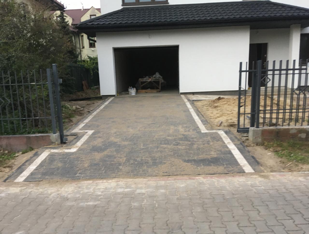 Nowo ułożony podjazd z szarej kostki brukowej z białymi obramowaniami, prowadzący do garażu, widoczny fragment ogrodzenia metalowego i przygotowane podłoże pod nasadzenia.