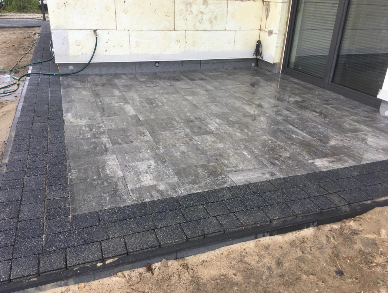 Wyłożony szarym brukiem taras z ciemną obwódką, przylegający do budynku z widocznymi elementami izolacji termicznej. W tle widoczne okno tarasowe i fragment trawnika.