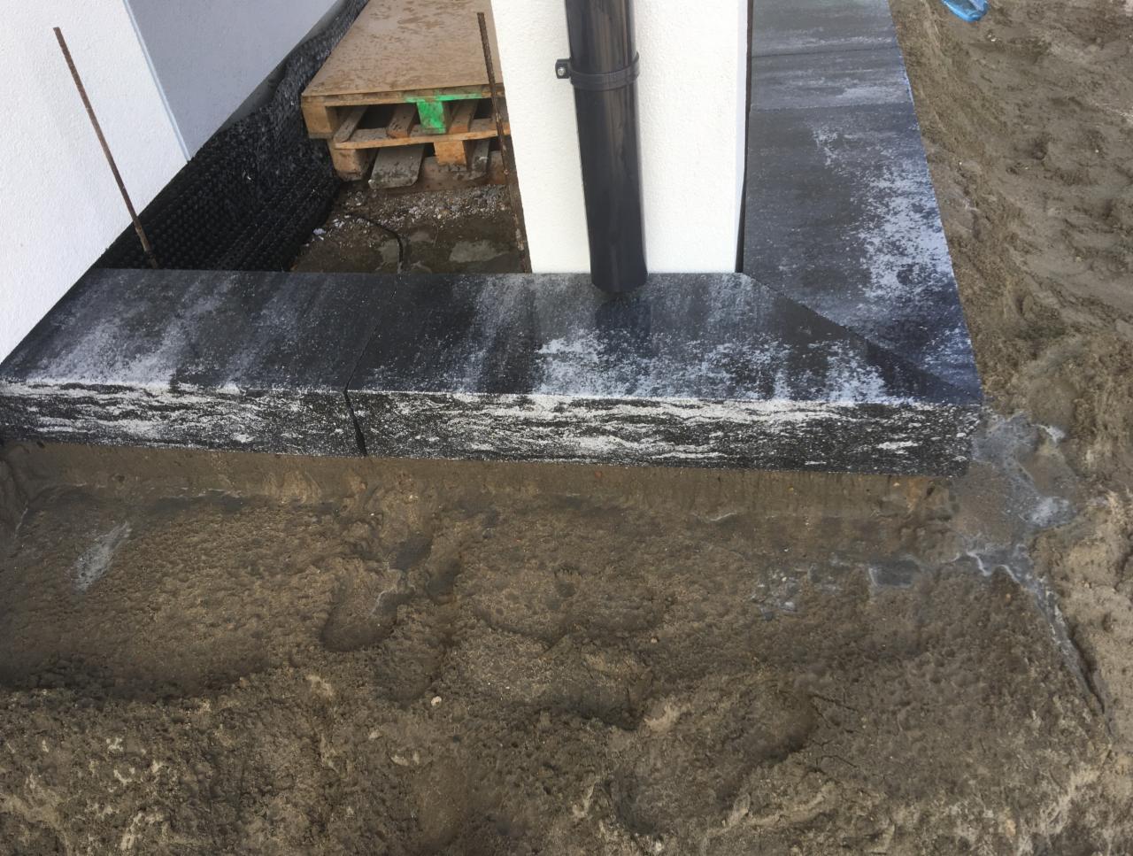 Fragment obłożonej czarną kostką granitową ściany budynku z widoczną rurą spustową, paletą w tle i ziemią w stanie surowym, ukazujący etap prac wykończeniowych.