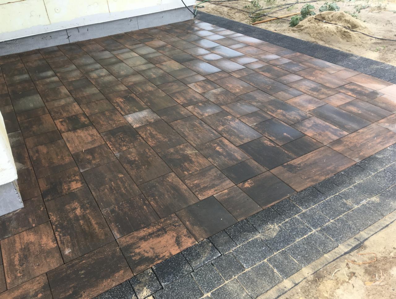 Ułożona kostka brukowa w odcieniach brązu i czerni, z czarnym obramowaniem, częściowo mokra, przylegająca do ściany budynku.
