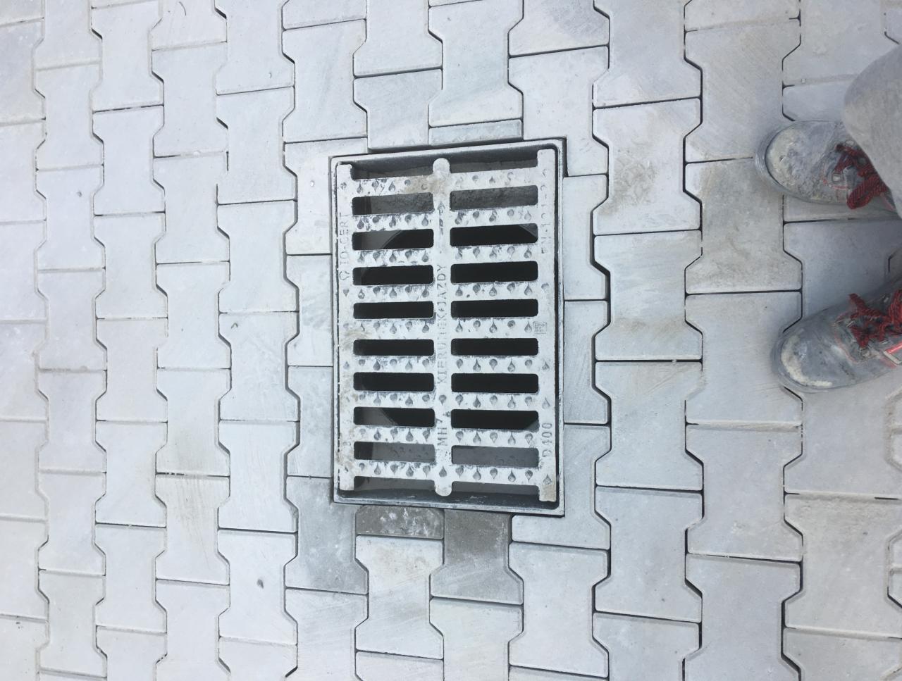 Ułożona szara kostka brukowa typu 'H' z metalową kratką ściekową w centralnej części; widoczne fragmenty obuwia roboczego z czerwonymi sznurówkami.
