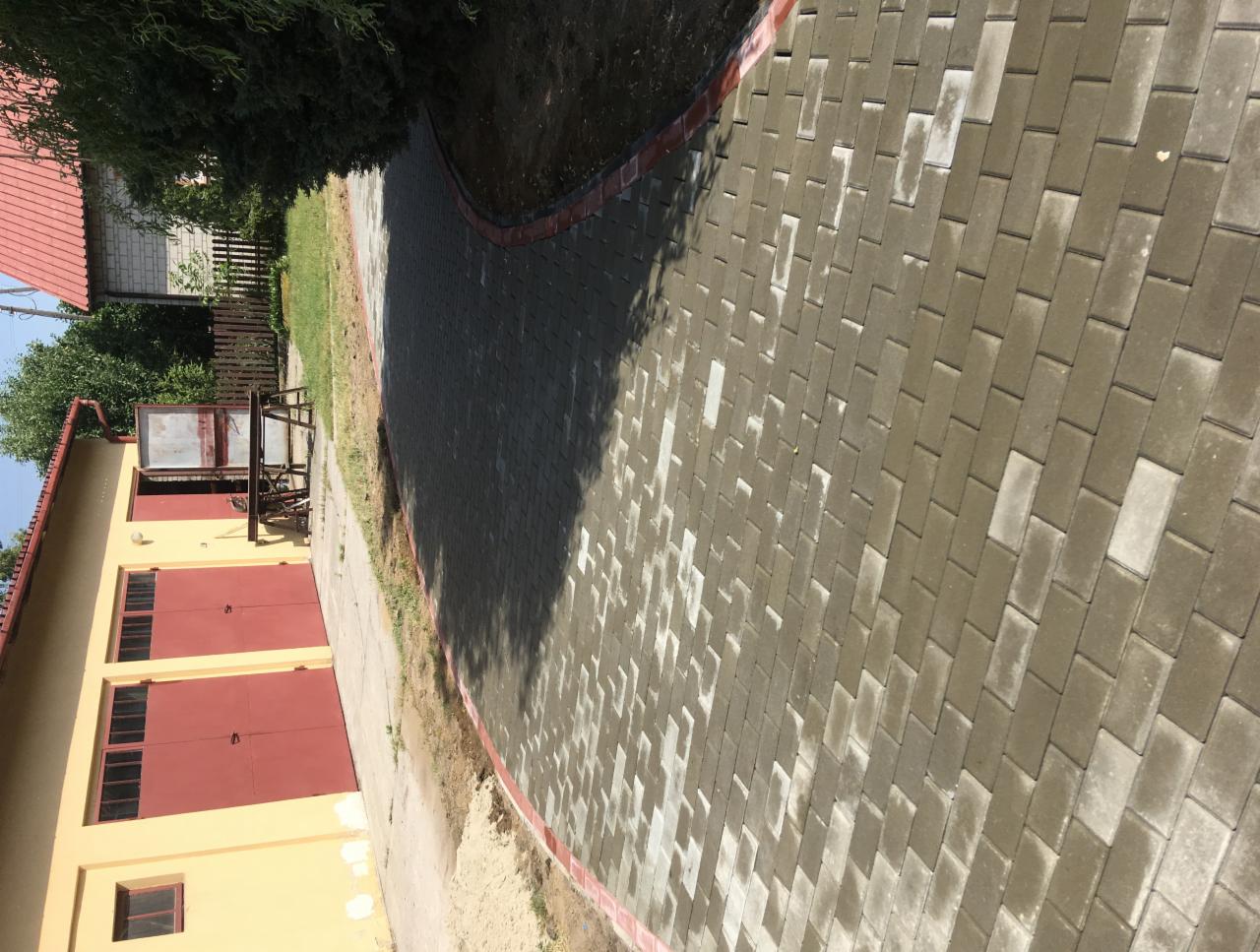 Zakrzywiony chodnik z szarej kostki brukowej z czerwoną obwódką, prowadzący obok budynku z czerwonymi drzwiami garażowymi i drzewa.