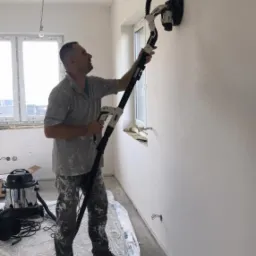 Pracownik szlifuje ścianę w jasnym pokoju za pomocą szlifierki z odkurzaczem, widoczne przygotowania do prac wykończeniowych i zabezpieczona folią podłoga.