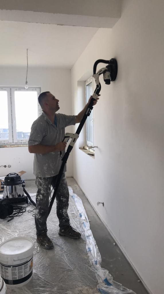 Pracownik szlifuje ścianę w jasnym pokoju za pomocą szlifierki z odkurzaczem, widoczne przygotowania do prac wykończeniowych i zabezpieczona folią podłoga.