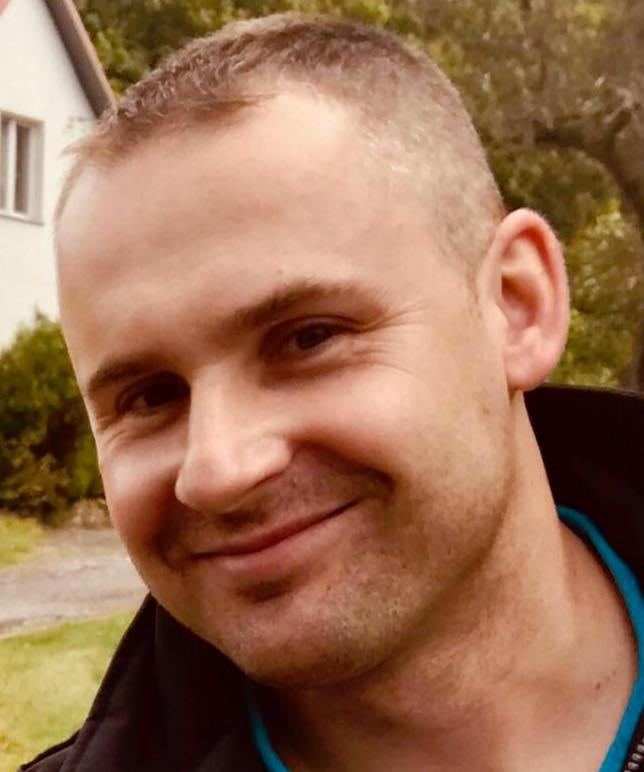 Uśmiechnięty mężczyzna z krótkimi włosami, ubrany w ciemną kurtkę z niebieską podszewką, na tle zieleni i fragmentu budynku.