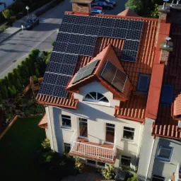 Widok z góry na dom z czerwoną dachówką i panelami słonecznymi na dachu, widoczna ulica i zieleń w ogrodzie.