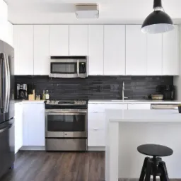 Nowoczesna, jasna kuchnia z białymi szafkami, czarnym backsplashiem i stalowymi urządzeniami, oświetlona wiszącymi lampami i naturalnym światłem.