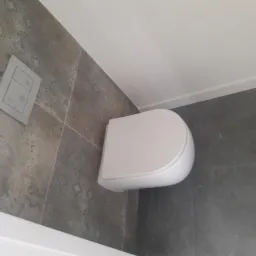 Nowoczesna toaleta podwieszana z szarą płytką dekoracyjną na ścianie i podłodze, widoczny przycisk spłukiwania.