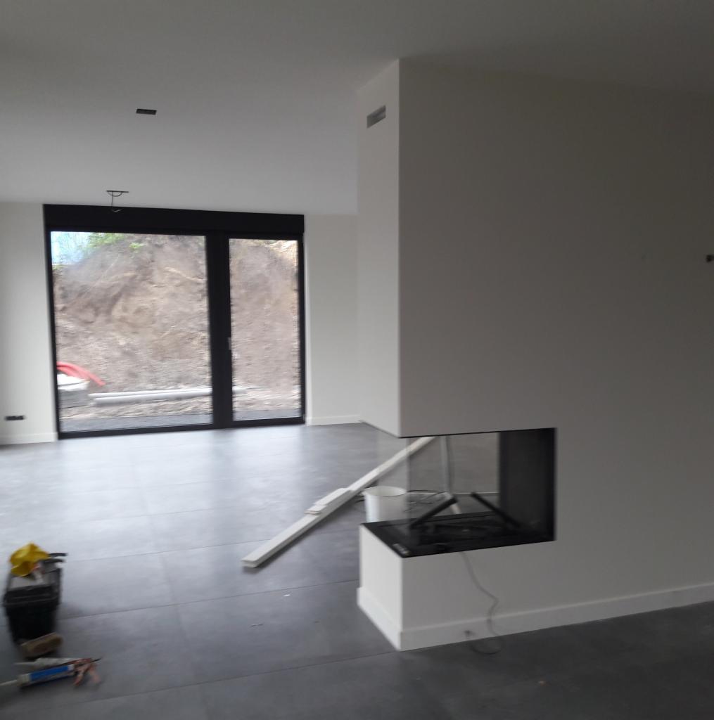 Wnętrze w trakcie wykańczania: szare płytki podłogowe, nowoczesny kominek z przeszklonym paleniskiem, duże okno tarasowe z widokiem na ziemny nasyp, narzędzia glazurnicze w rogu pomieszczenia.