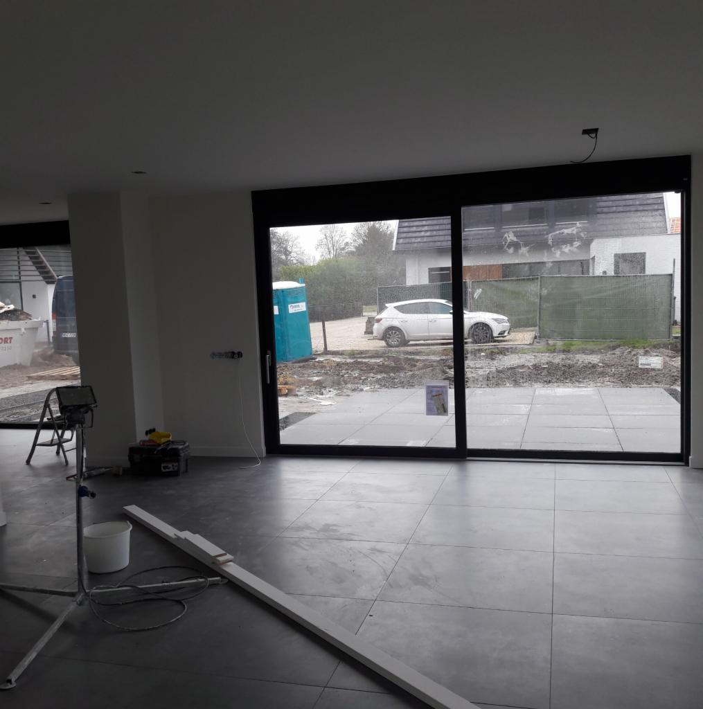 Wnętrze domu w trakcie wykańczania, widoczne szare płytki podłogowe, duże okno przesuwne z widokiem na posesję z kostką, samochodem i przenośną toaletą, narzędzia glazurnicze w tle.