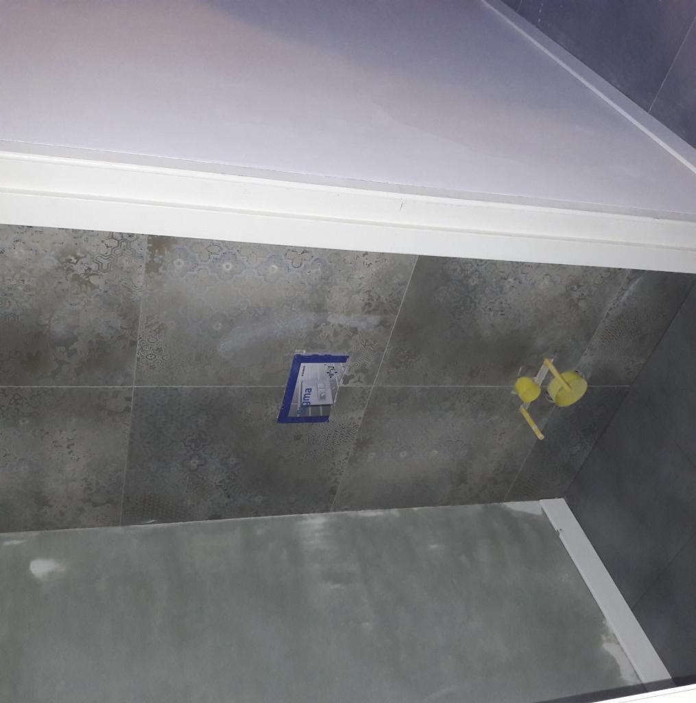 Świeżo ułożone, szare, wzorzyste płytki ceramiczne na ścianie łazienki, widoczny fragment przygotowanej do malowania ściany oraz żółty element instalacji wodnej.