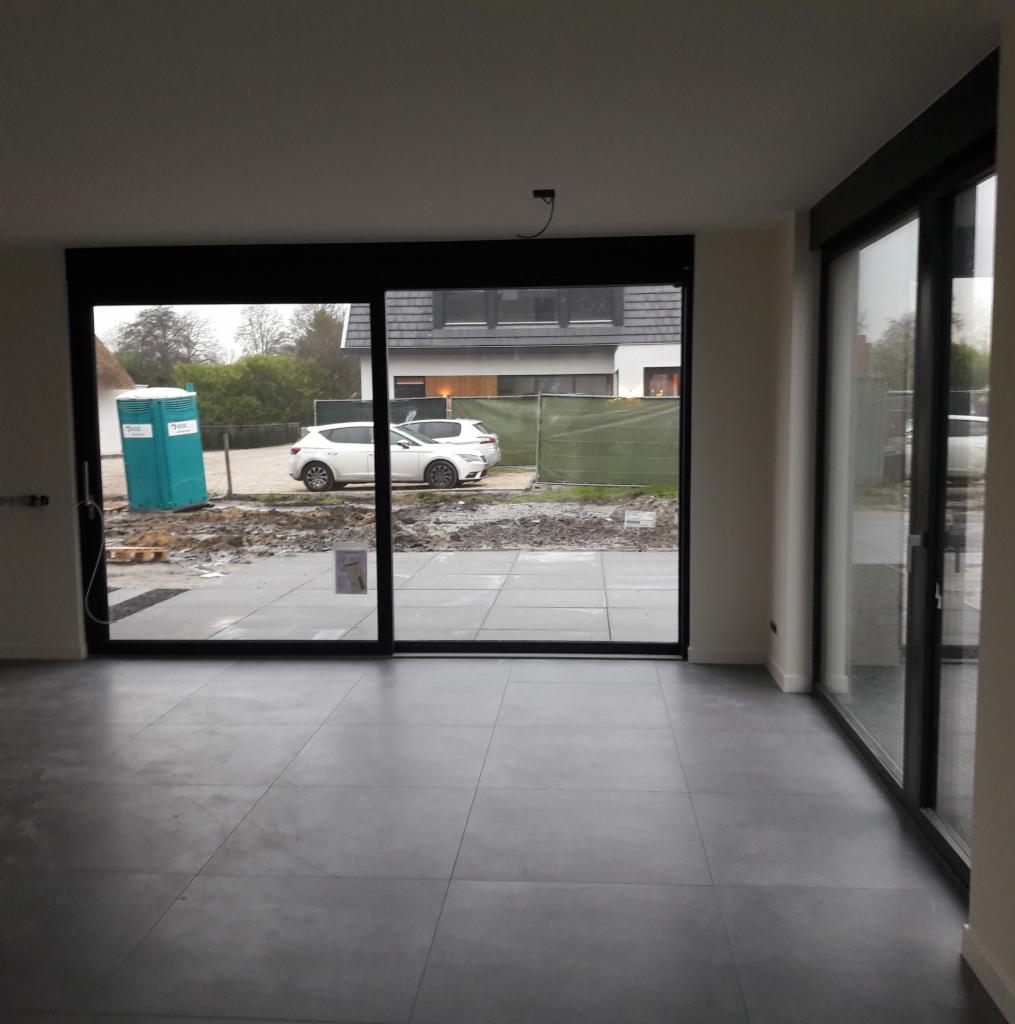 Wnętrze nowego domu z szarymi, kwadratowymi płytkami podłogowymi, widok na plac budowy przez duże okno tarasowe.