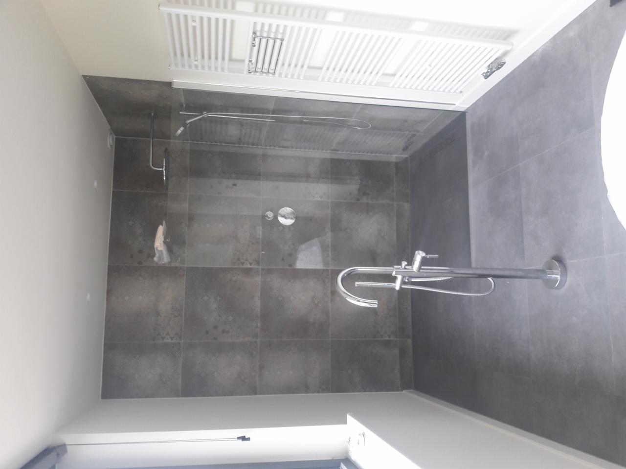 Szara kabina prysznicowa z deszczownicą i baterią termostatyczną, wykończona kwadratowymi płytkami z subtelnym wzorem, widoczny biały grzejnik drabinkowy na suficie.