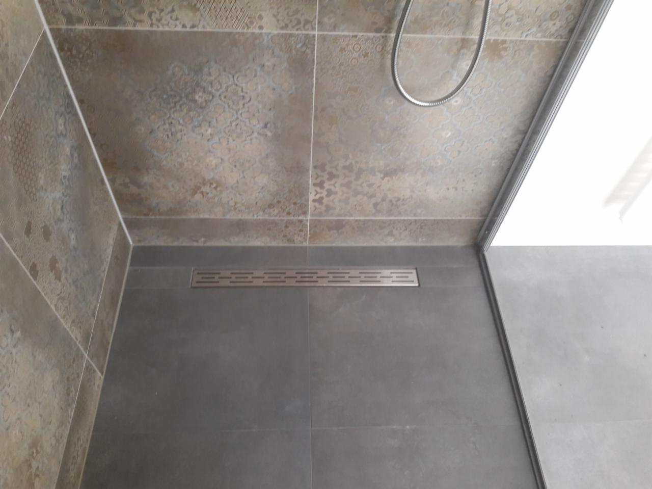 Wykończona łazienka z szarymi płytkami podłogowymi i dekoracyjnymi płytkami ściennymi w odcieniach beżu i brązu, widoczny liniowy odpływ prysznicowy i fragment słuchawki prysznicowej.
