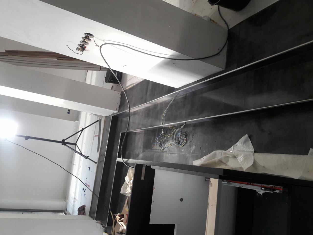 Montaż czarnej, błyszczącej glazury w wąskiej wnęce, widoczne przewody elektryczne i lampa budowlana na statywie.