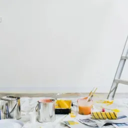 Przygotowania do malowania ścian: wałek oparty o ścianę, aluminiowa drabina, pędzle w wiaderku z farbą, puszki z farbą, kuweta malarska i gazety zabezpieczające podłogę.