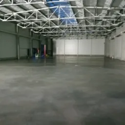 Gładka, szara posadzka w przestronnym, pustym magazynie z metalową konstrukcją dachu i jasnymi ścianami.