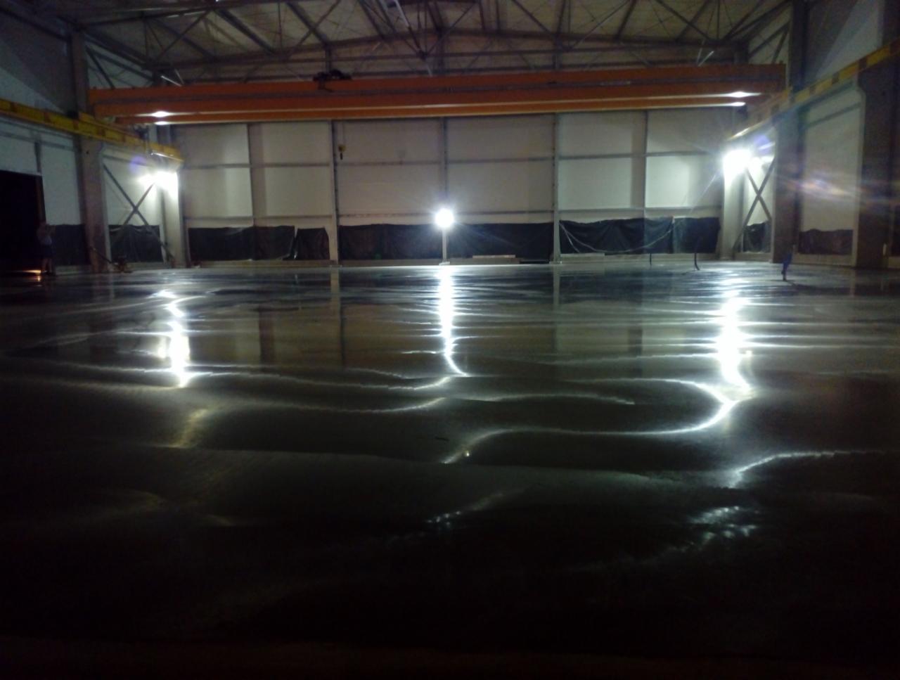 Świeżo wylana, mokra posadzka w hali przemysłowej, odbijająca światła lamp sufitowych i dźwigu pomostowego.