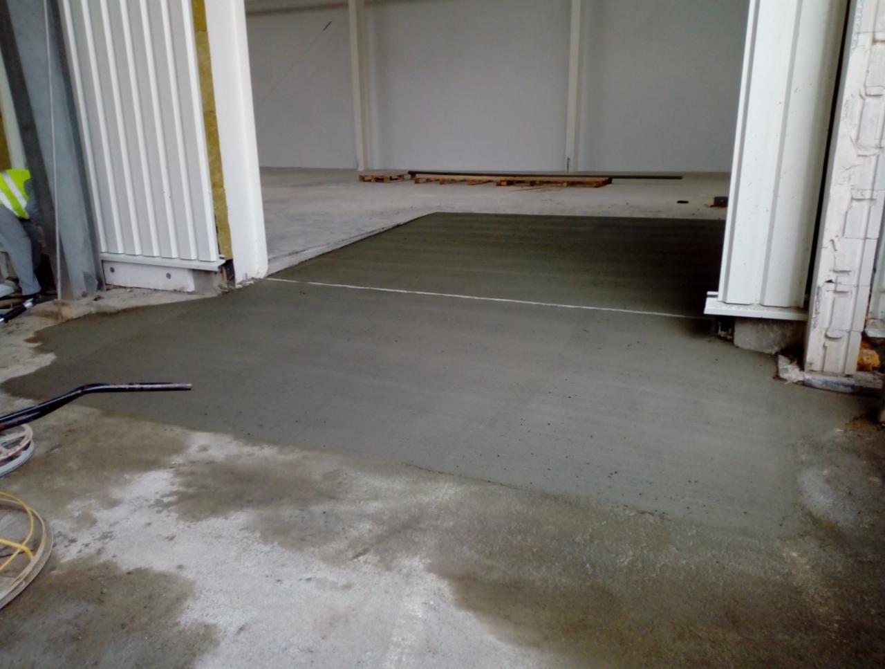 Świeżo wylana, gładka posadzka przemysłowa w hali magazynowej, z widocznym narzędziem do rozprowadzania betonu i fragmentem pracownika w kamizelce odblaskowej.