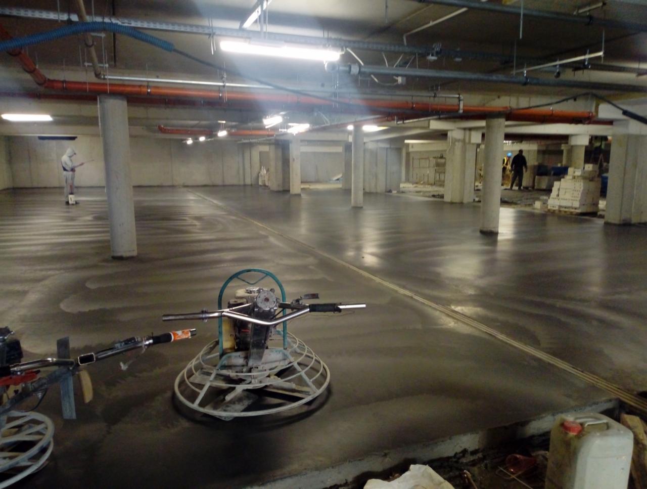 Świeżo wylana, gładka betonowa posadzka w hali garażowej, zacieraczki mechaniczne w użyciu, pracownik w kombinezonie ochronnym w tle.