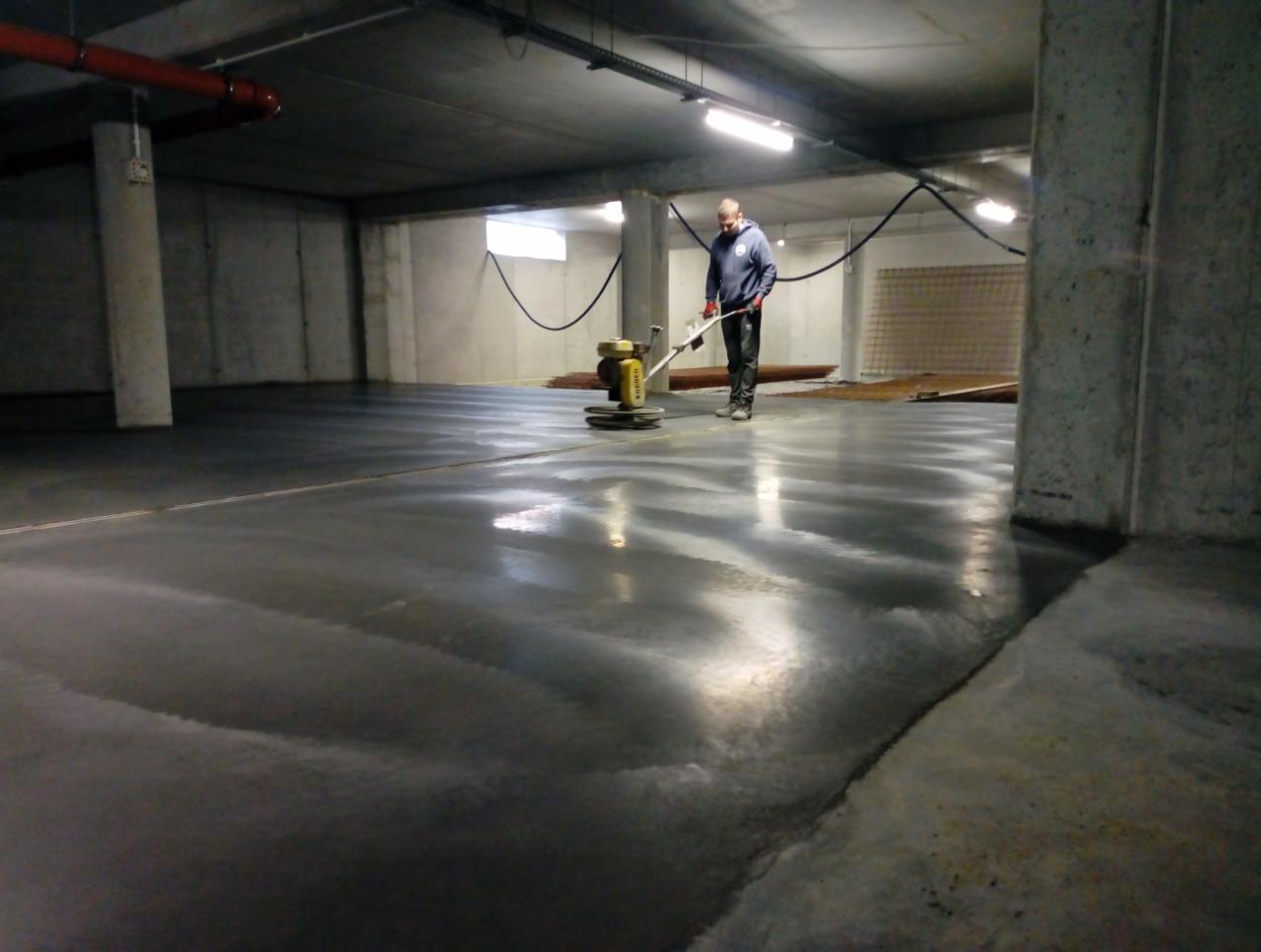 Pracownik w niebieskiej bluzie szlifuje mokrą, betonową podłogę hali przemysłowej za pomocą maszyny polerskiej; widoczne ślady po polerowaniu i kolumna konstrukcyjna w tle.