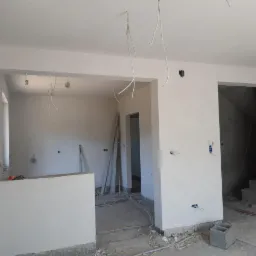 Wnętrze w trakcie prac wykończeniowych; świeżo nałożone gładzie na ścianach, widoczne wystające kable elektryczne, drabiny oparta o ścianę w głębi pomieszczenia, surowy beton na podłodze.