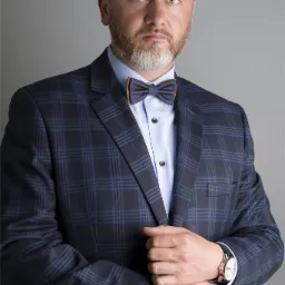 Portret mężczyzny w granatowym garniturze w kratę, z drewnianą muchą i zegarkiem na ręce, na jednolitym szarym tle.