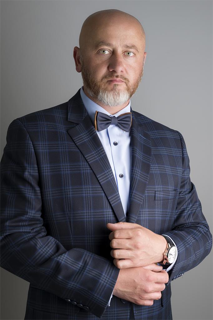 Portret mężczyzny w granatowym garniturze w kratę, z drewnianą muchą i zegarkiem na ręce, na jednolitym szarym tle.