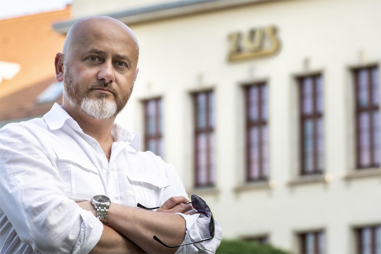 Portret mężczyzny z brodą w białej koszuli, z zegarkiem na ręku i okularami przeciwsłonecznymi, na tle budynku ZUS.